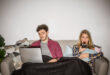 a guy sitting on a couch and using laptop and a girl with him watching tv